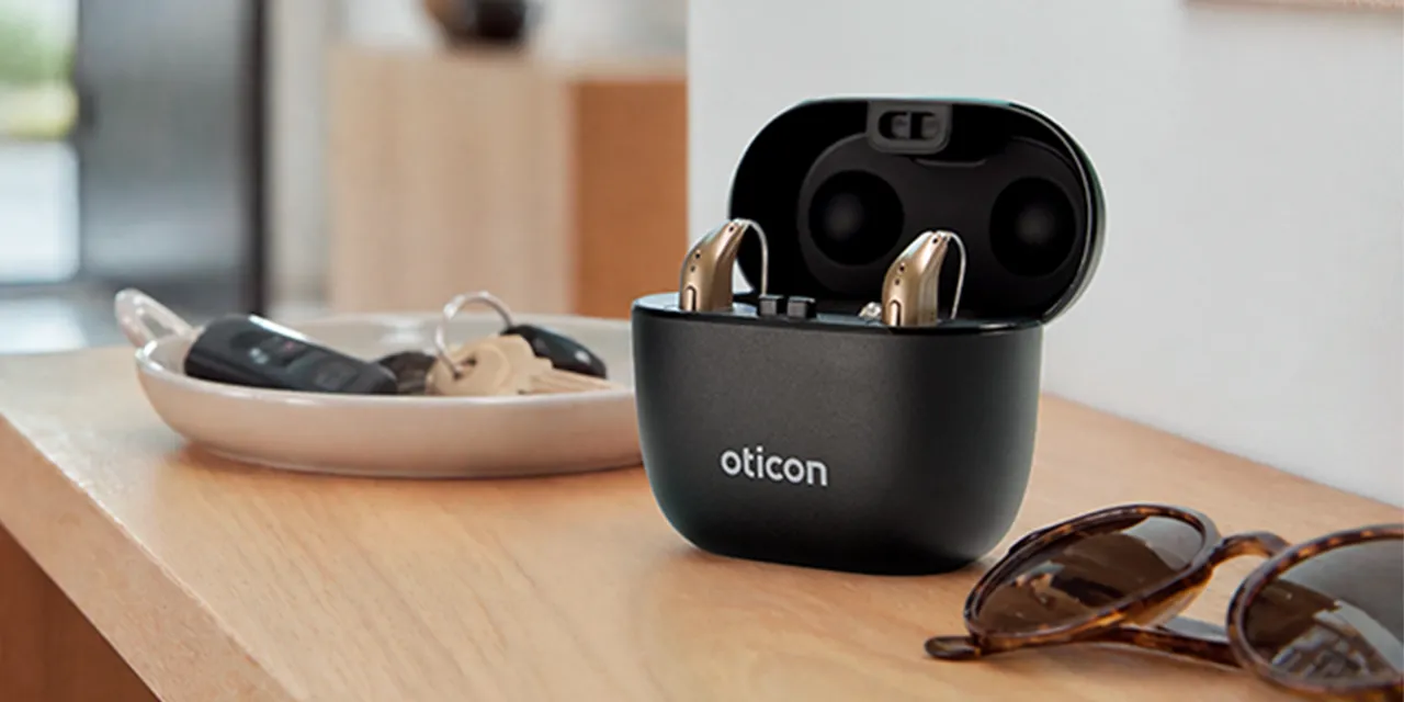 The Oticon SmartCharger case open on a wooden desk with a pair of Oticon Reveal hearing aids inside
